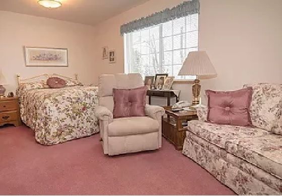 A cozy living room with traditional decor, featuring armchairs and a floral-patterned sofa on pink carpeting.