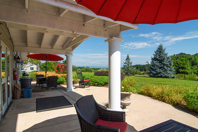 A charming patio with red umbrellas offers a scenic view of a landscaped garden and distant trees on a sunny day.