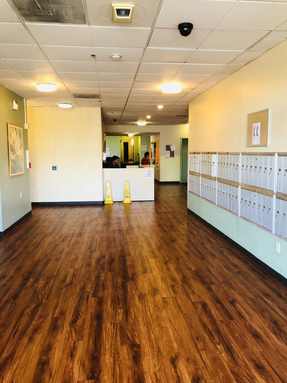 A hallway in an office building with wooden flooring, mailboxes on the right, and a reception area ahead.