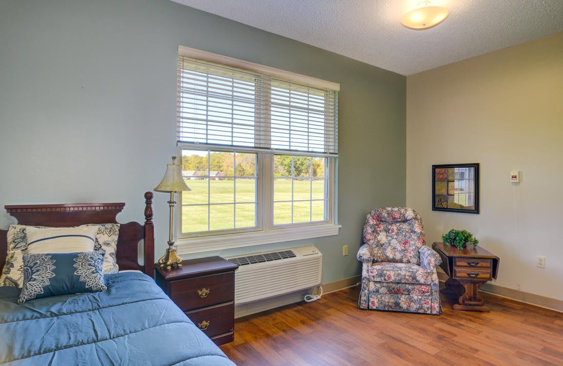 A cozy bedroom with wooden flooring, featuring a bed, nightstand, lamp, and a floral armchair by the window.
