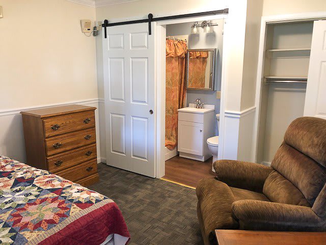 A cozy bedroom with a barn door leading to an adjacent bathroom, featuring a bed, wooden dresser, and armchair.