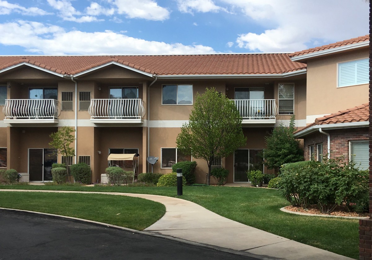 A two-story residential building with balconies, surrounded by trees and landscaping under a partly cloudy sky.