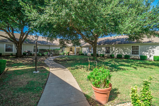 A serene courtyard features a large tree, garden landscaping, and a concrete pathway leading to residential buildings.