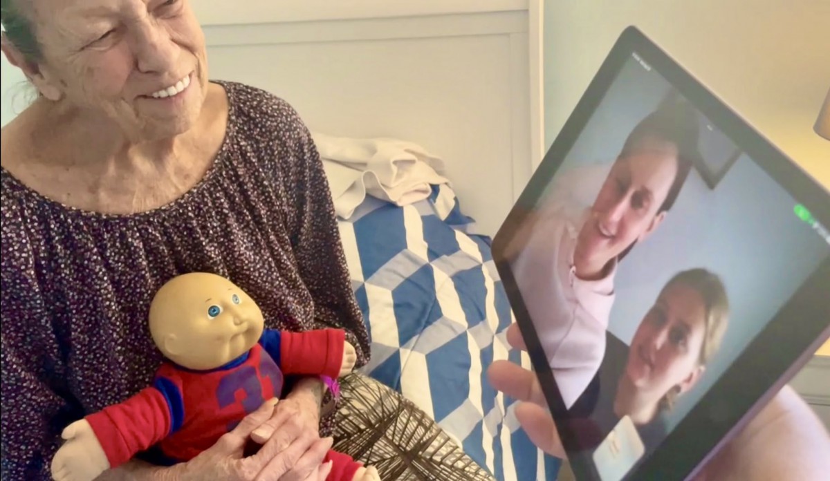 A person holding a doll is on a video call with two people displayed on a laptop screen in a bedroom setting.