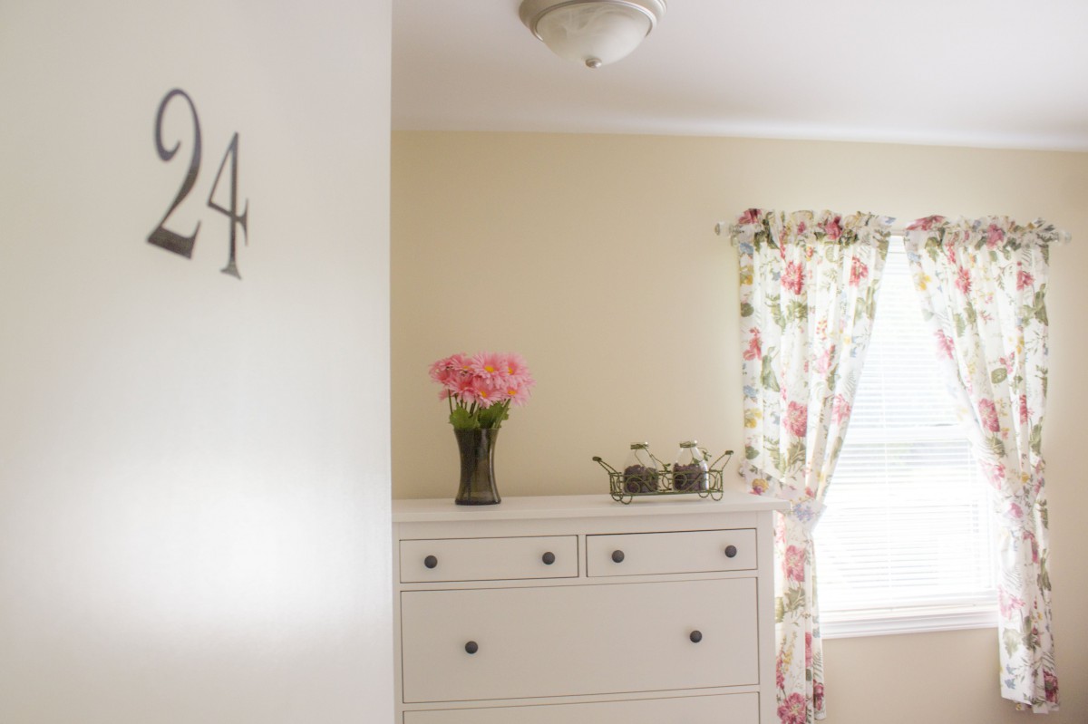A cozy bedroom with a white dresser, floral curtains, and a vase of pink flowers.