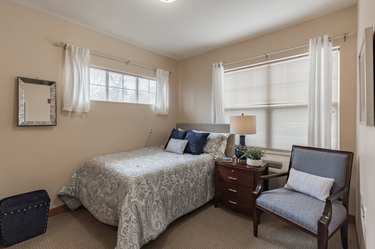 A cozy bedroom with a bed, nightstand, lamp, and chair by the windows.