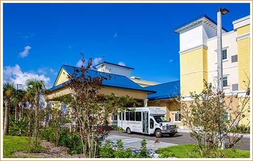 A yellow building with a blue roof and a white bus parked in front, surrounded by trees under a clear blue sky.