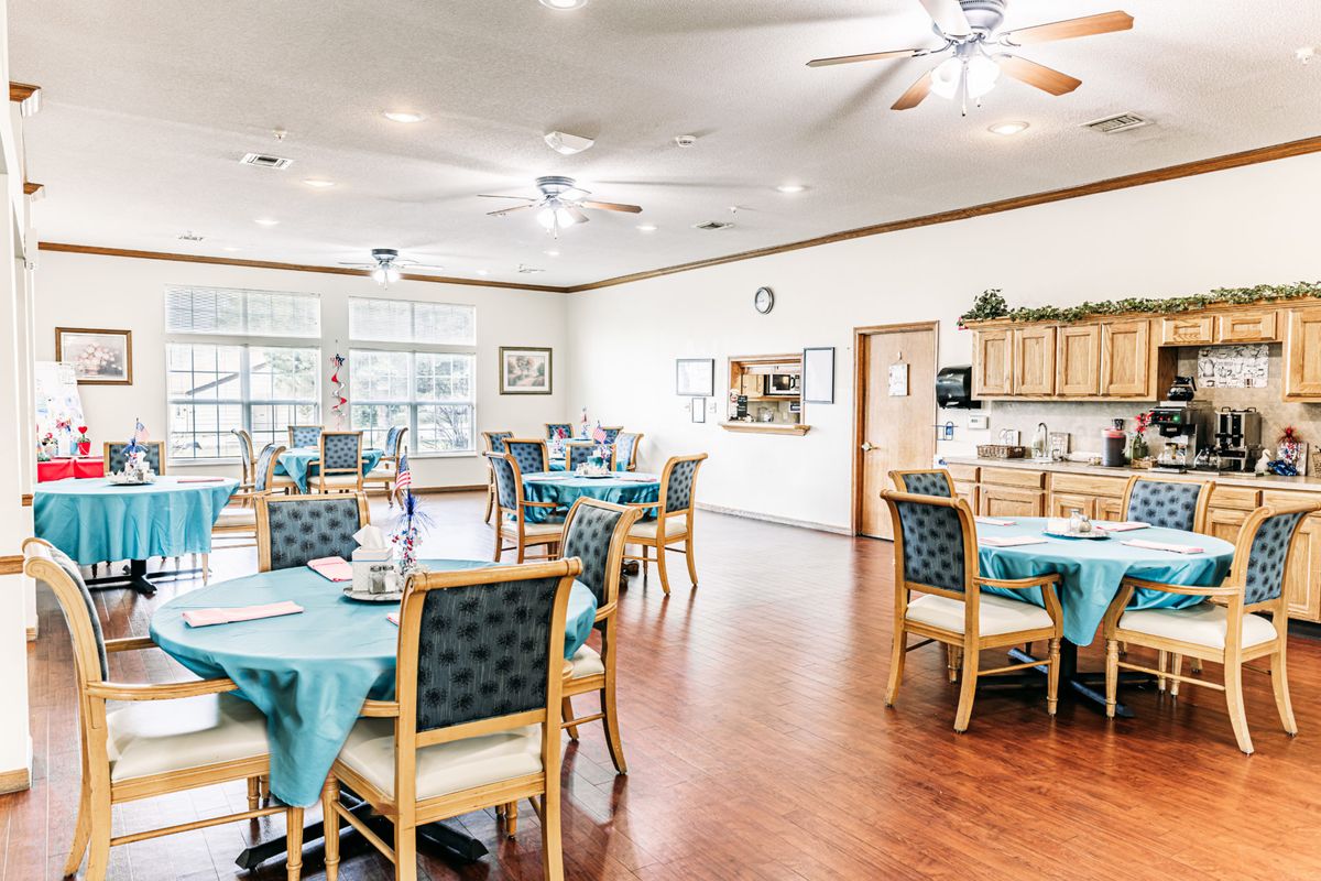 A spacious and well-lit dining area with neatly arranged tables and chairs, featuring a kitchen area in the background.