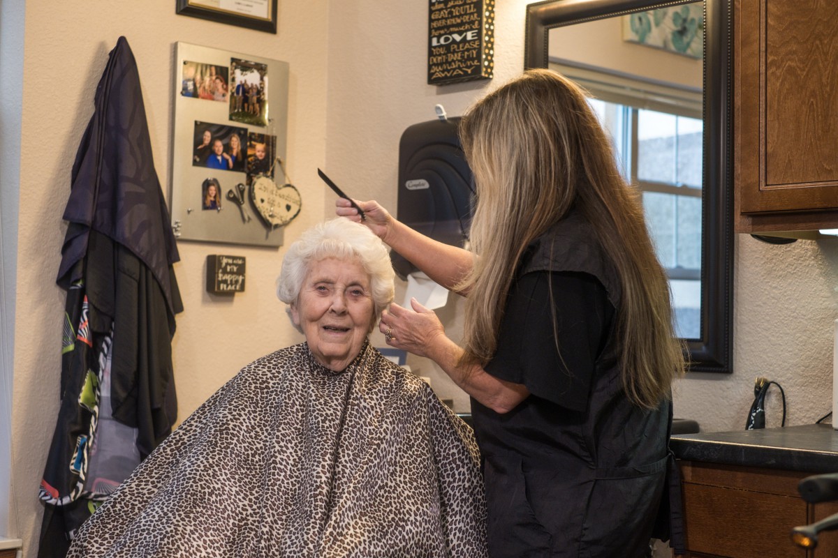 An elderly woman receives a haircut from a hairdresser in a cozy salon setting, surrounded by personal photos and decor.