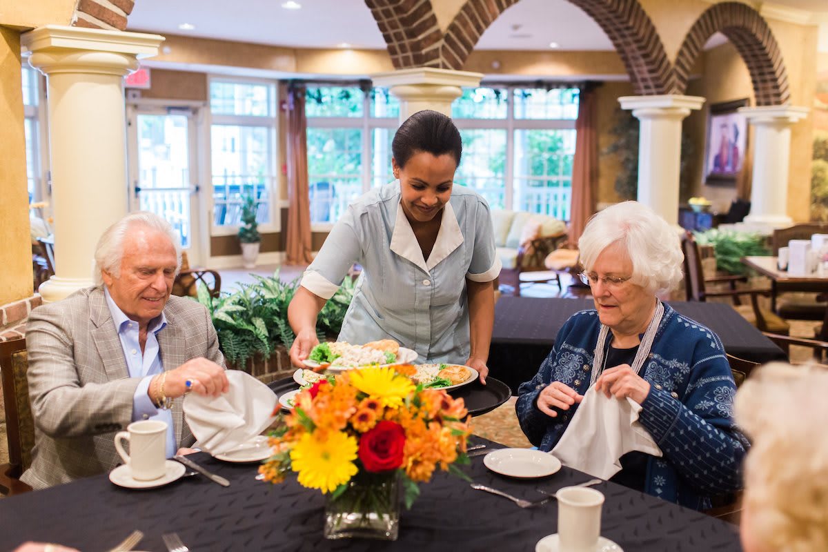 A waitress serves an elderly couple in a restaurant setting, with a colorful floral arrangement on the table.