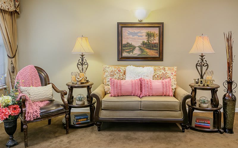 A cozy living room with a sofa adorned with pink pillows, flanked by decorative lamps and side tables, and a landscape painting on the wall.