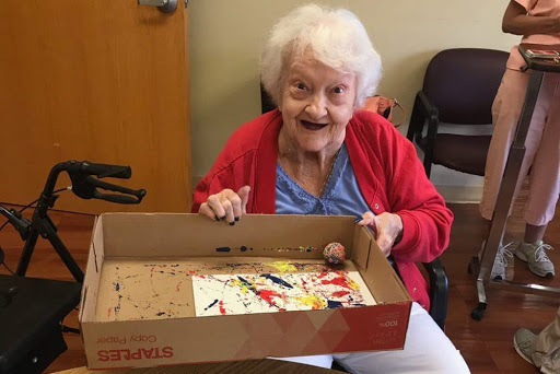 An elderly woman joyfully displays her artwork after an engaging painting session.