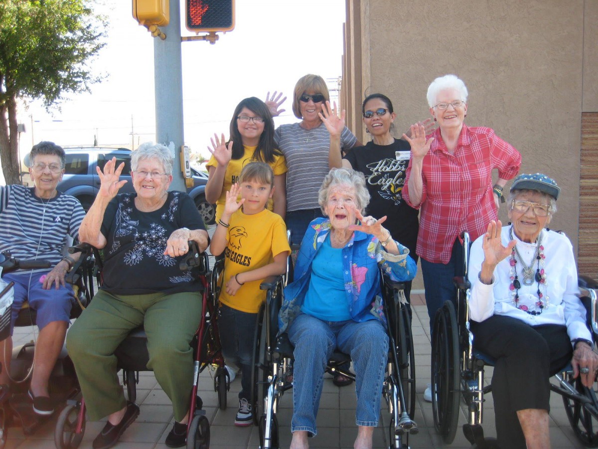 A group of people of various ages, some in wheelchairs, smiling and waving outdoors.