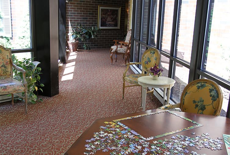 A cozy hallway with a puzzle on a table, flanked by elegant chairs and large windows letting in natural light.
