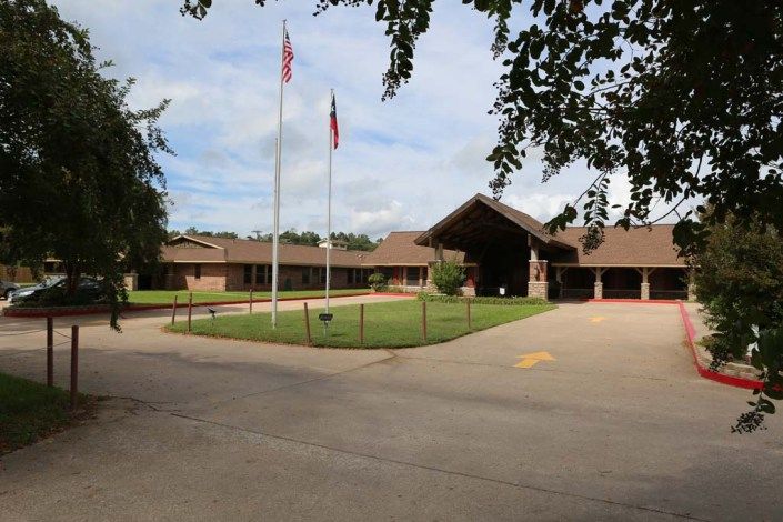 A single-story brick building with flags in front and a driveway, surrounded by greenery.
