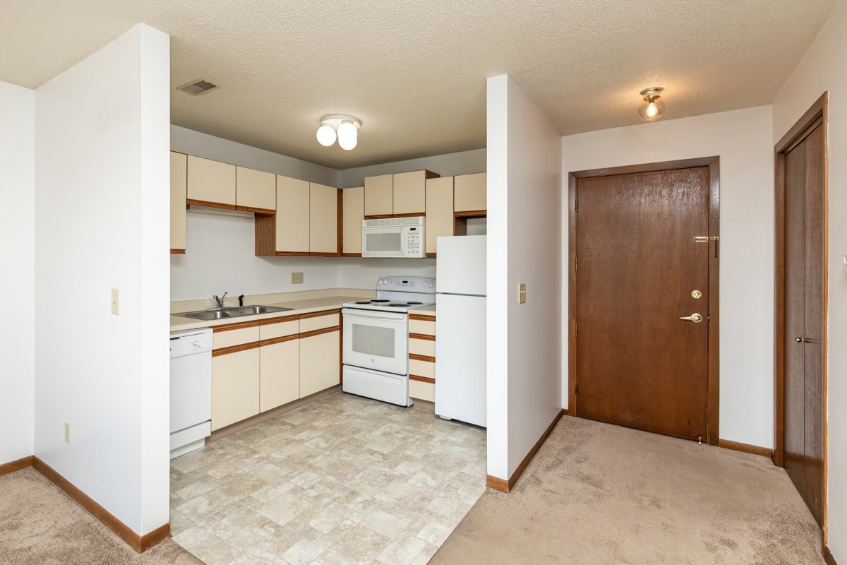 A small apartment kitchen with white cabinets, a stove, microwave, refrigerator, and sink next to an entrance door.