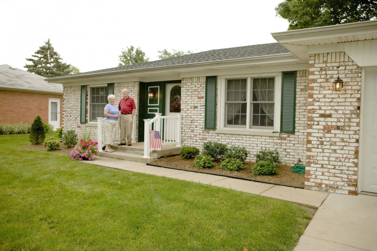 A classic brick house with a well-maintained garden and two people standing on the porch.