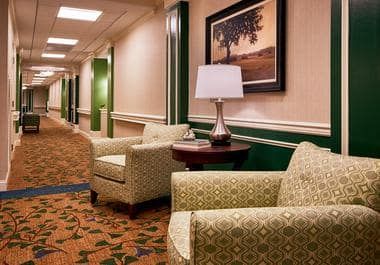 A cozy hotel lobby featuring comfortable armchairs, a lamp on a small table, and framed artwork along a carpeted hallway.
