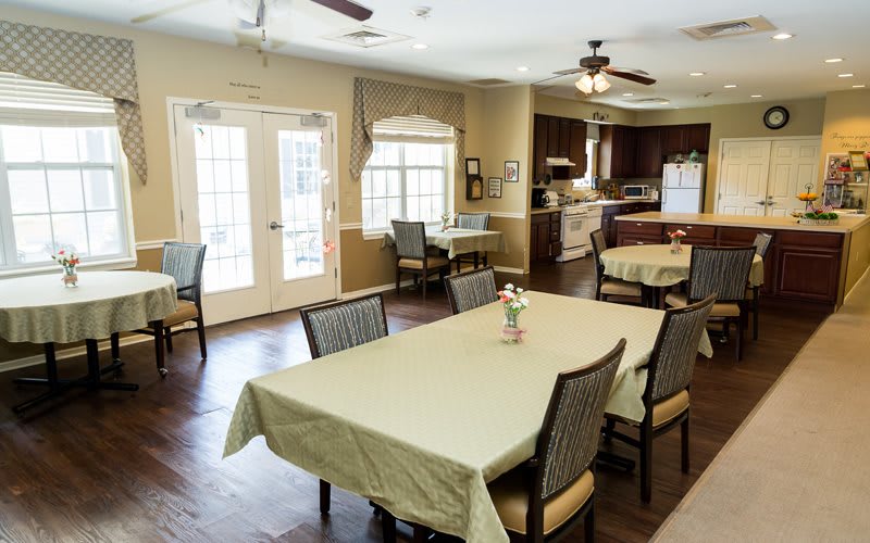 A cozy dining area with multiple tables and chairs, connected to a kitchen space, featuring large windows and ceiling fans.