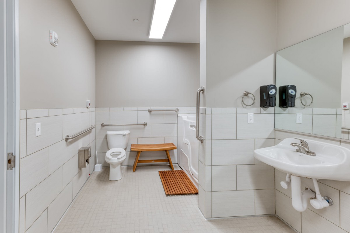 A clean and modern accessible bathroom with a toilet, sink, handrails, and a wooden bench.