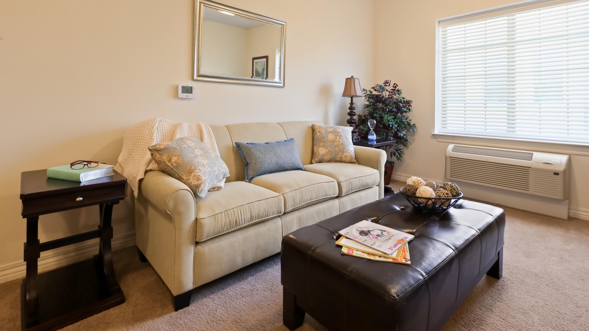 A cozy living room with a beige sofa, decorative cushions, and a dark coffee table adorned with magazines and a bowl. A mirror hangs above the sofa, and a lamp sits in the corner next to a plant.