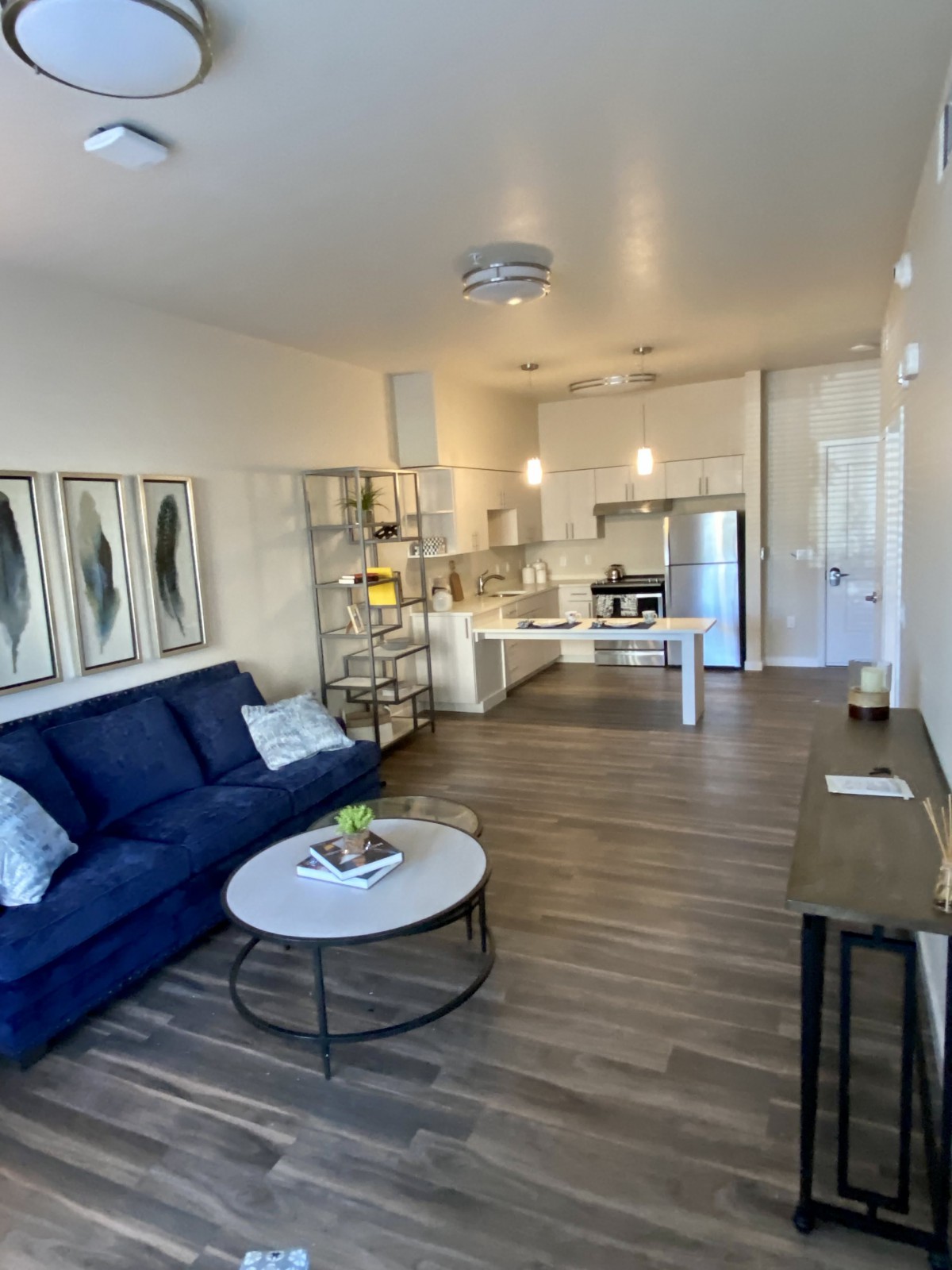 A modern living room with an open floor plan featuring a blue sofa, wood flooring, and a kitchen area.