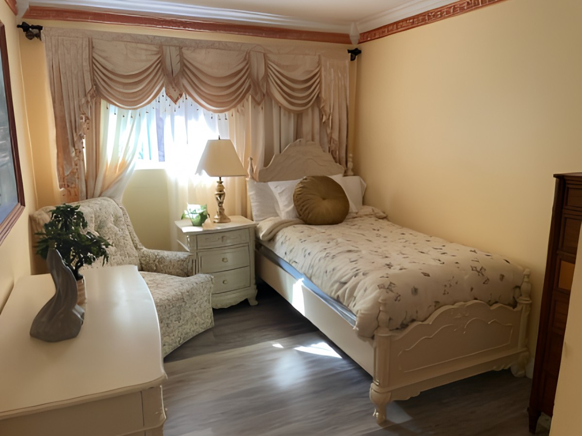 A cozy bedroom featuring a single bed with elegant bedding, an armchair, and a lamp by the window letting in natural light.