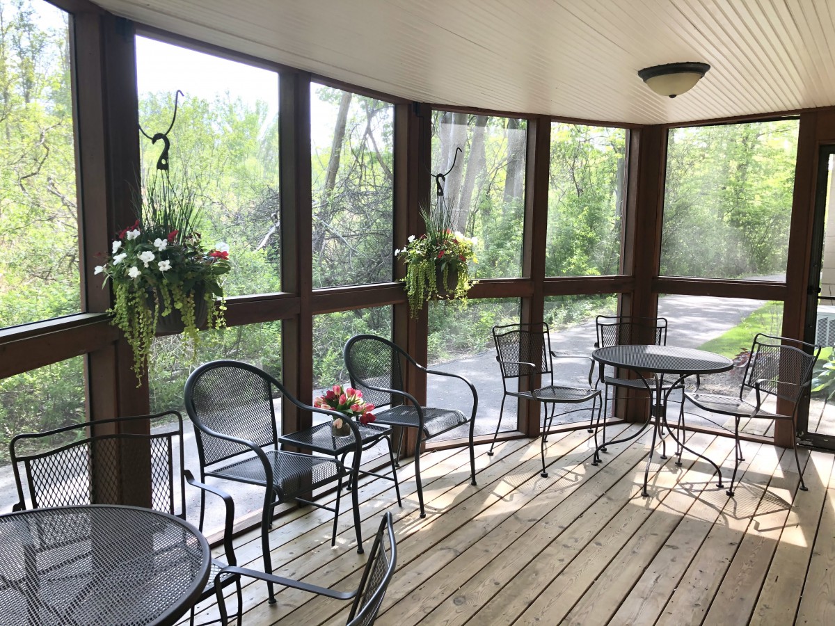 A cozy porch with tables and chairs, surrounded by windows and decorated with hanging plants.