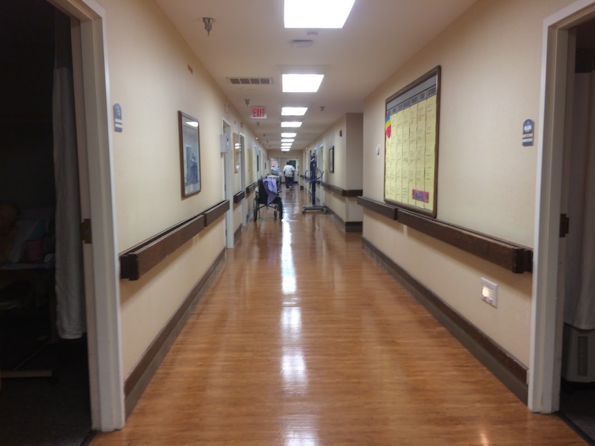 A long hospital corridor with wooden flooring and bright overhead lighting.