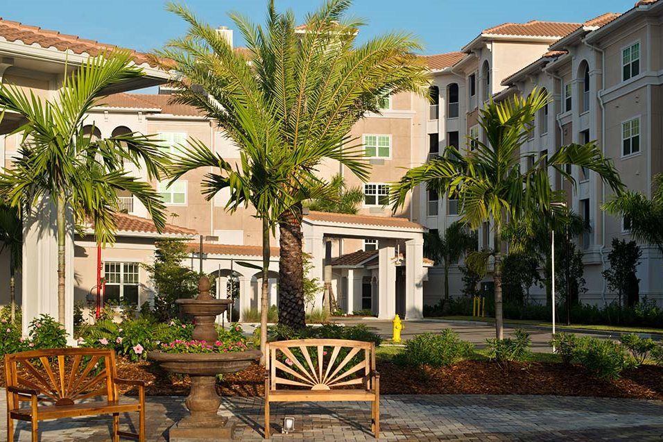 A scenic view of a building surrounded by palm trees and a garden with a wooden bench in the foreground.