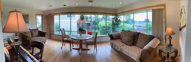 A bright dining and living area with large windows, allowing natural light to fill the space. The room features a round table with chairs, a comfortable sofa, and indoor plants on a hardwood floor.