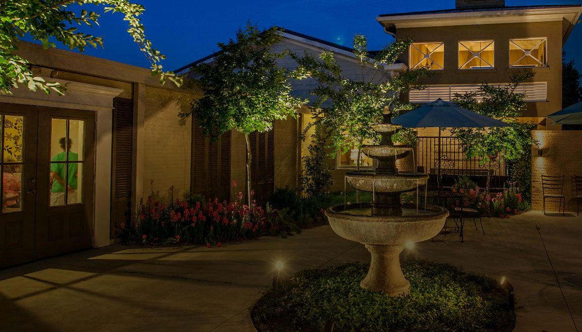 A serene nighttime courtyard featuring a lit fountain surrounded by trees and a building.