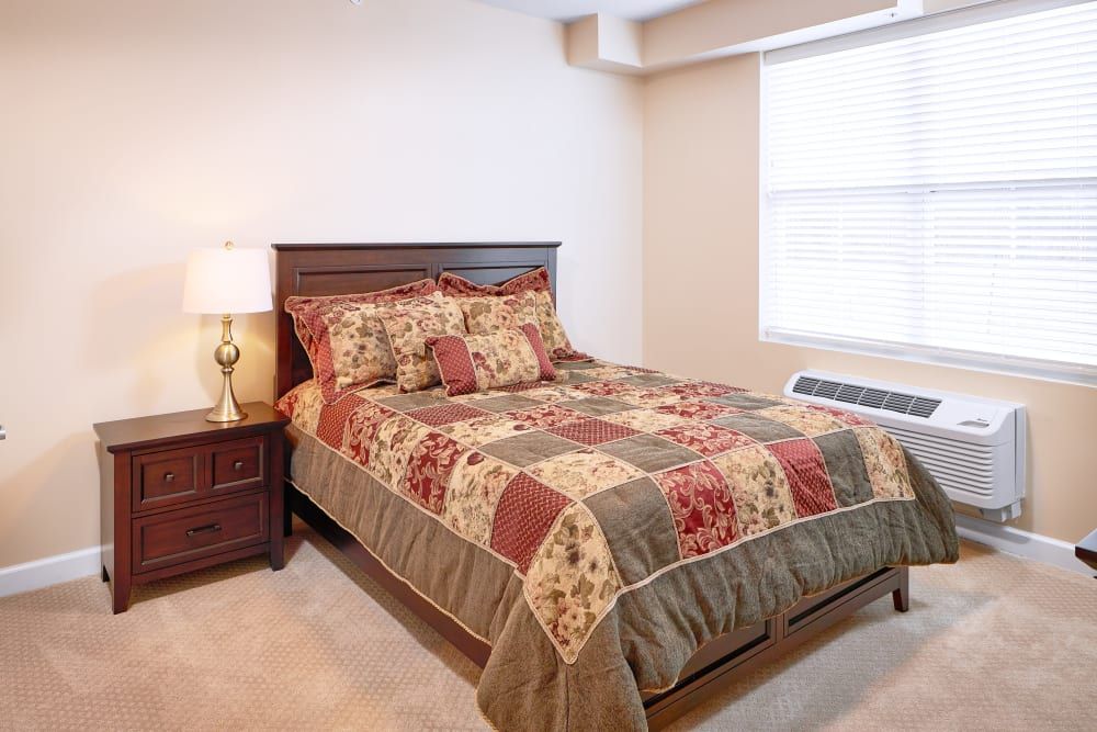 A cozy bedroom featuring a wooden bed with a patchwork quilt, a nightstand with a lamp, and natural light streaming through the window.