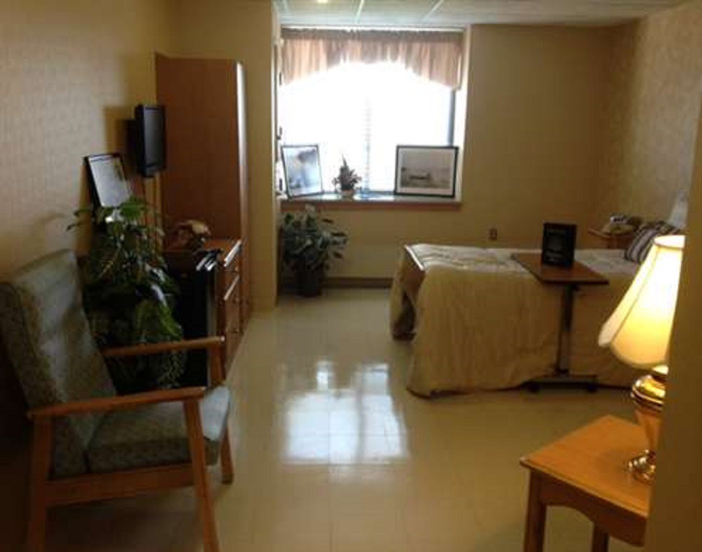 A tidy and well-lit hospital room with a bed, chair, and plants near the window.