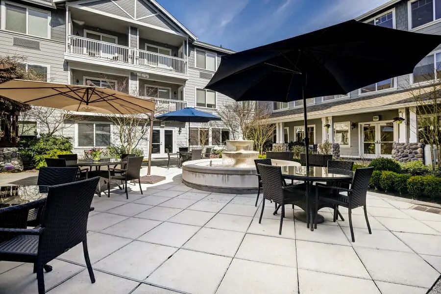 A modern outdoor courtyard featuring patio furniture, umbrellas, and a central fountain in front of a multi-story building.