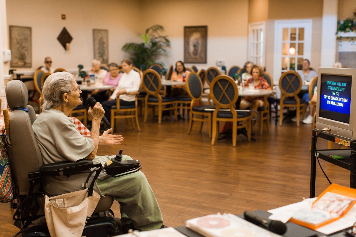 A group of seniors gather in a community room for social activities at a nursing home.