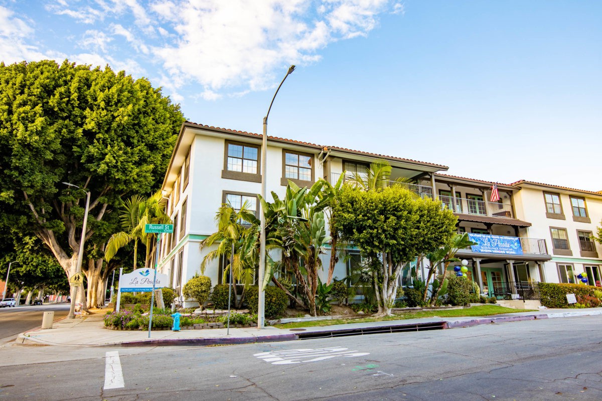 A residential apartment building on a sunny day, surrounded by trees and located at a street corner.