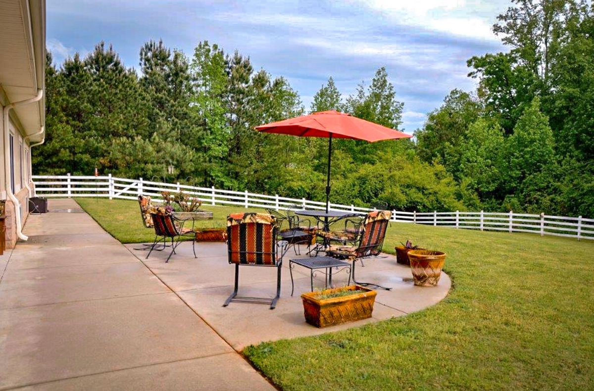 A serene backyard patio with outdoor furniture and a red umbrella, surrounded by greenery and a white fence.