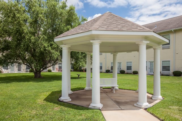 A white gazebo with multiple columns sits on a landscaped grassy area under a blue sky.