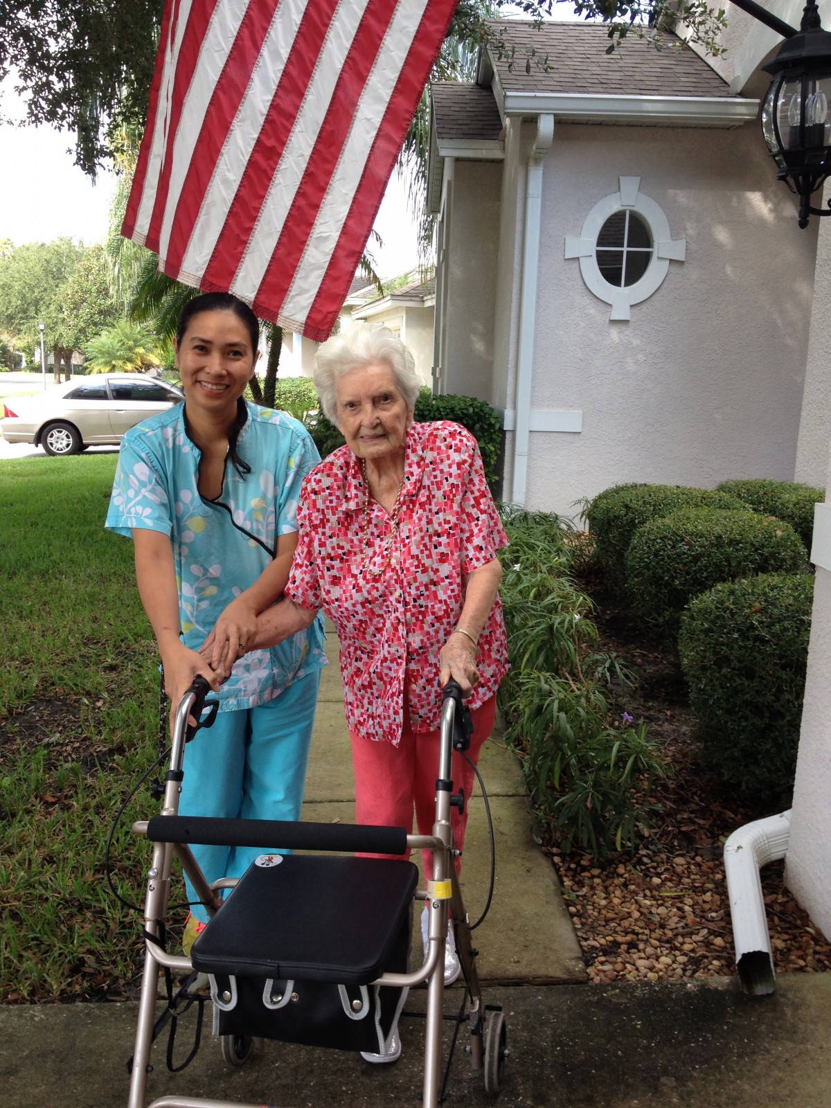 An elderly woman with a walker stands next to a caregiver outside a house, with an American flag in the background.