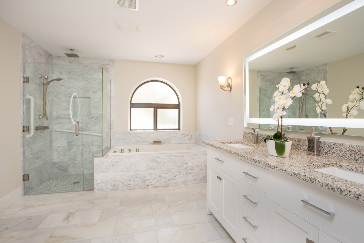 A modern luxury bathroom with marble finishes, featuring a large vanity, illuminated mirror, spacious shower, and bathtub with natural light streaming through an arched window.