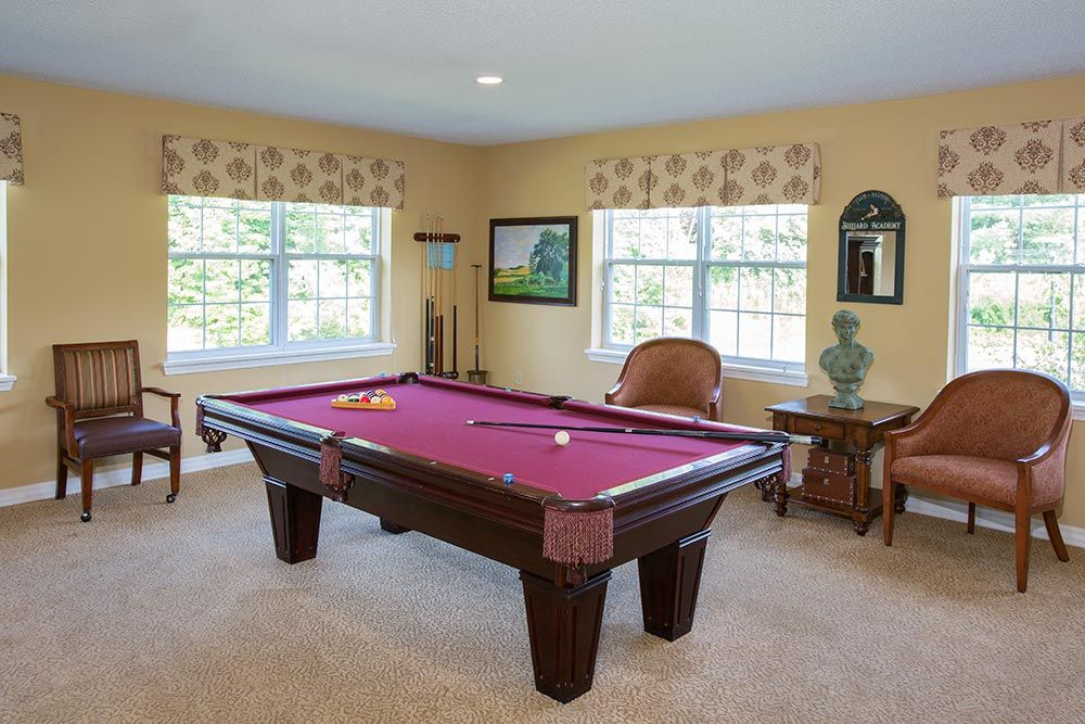 A well-lit game room featuring a billiards table with magenta felt, surrounded by windows and decorated with artwork and furniture.