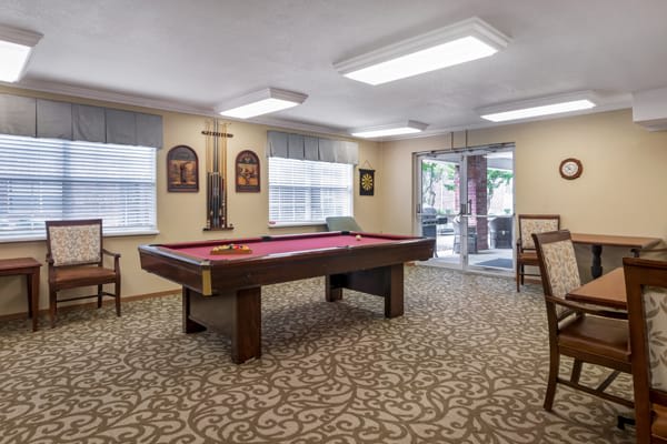 A game room featuring a pool table in the center, surrounded by chairs and natural lighting from large windows.