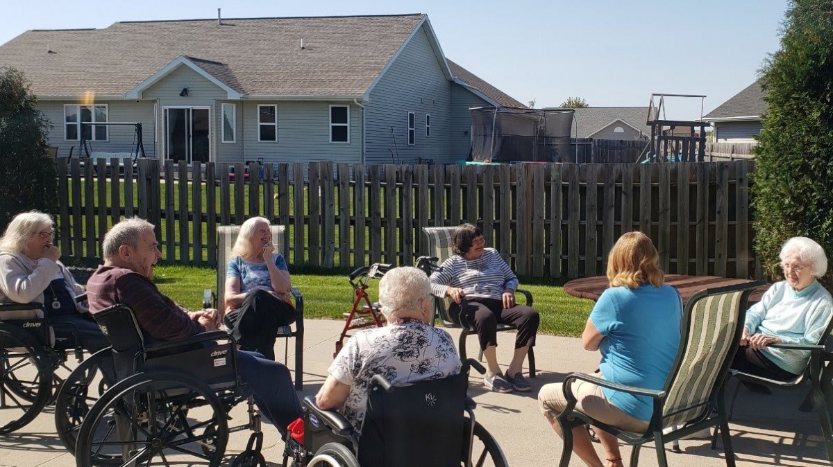 A group of elderly people sitting outdoors, some in wheelchairs, enjoying a sunny day in a garden area.