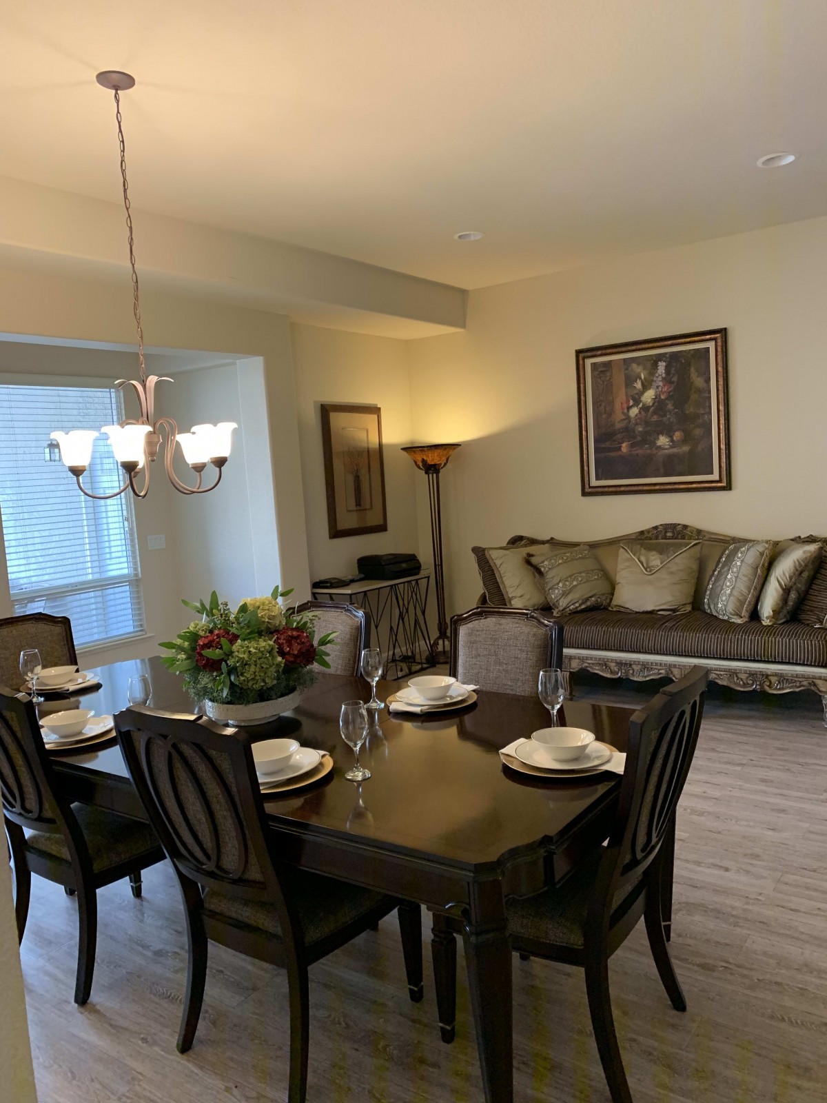 A cozy dining and living area featuring a dining table set for four, a comfortable sofa, elegant artwork, and a chandelier.