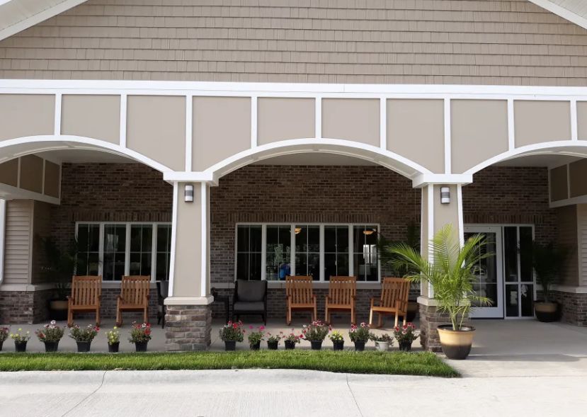 An outdoor porch area featuring several wooden chairs and potted plants in front of a building with arched details.