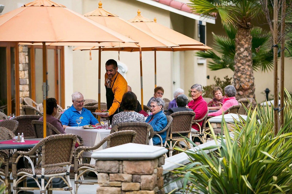 A group of seniors enjoying a meal outdoors at a restaurant patio, attended by a waiter.