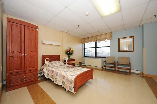 A well-lit hospital room featuring a bed, seating area, and decorative flowers.