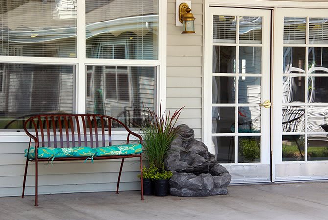 A cozy porch setup with a bench, cushion, plants, and a decorative rock next to a glass door.