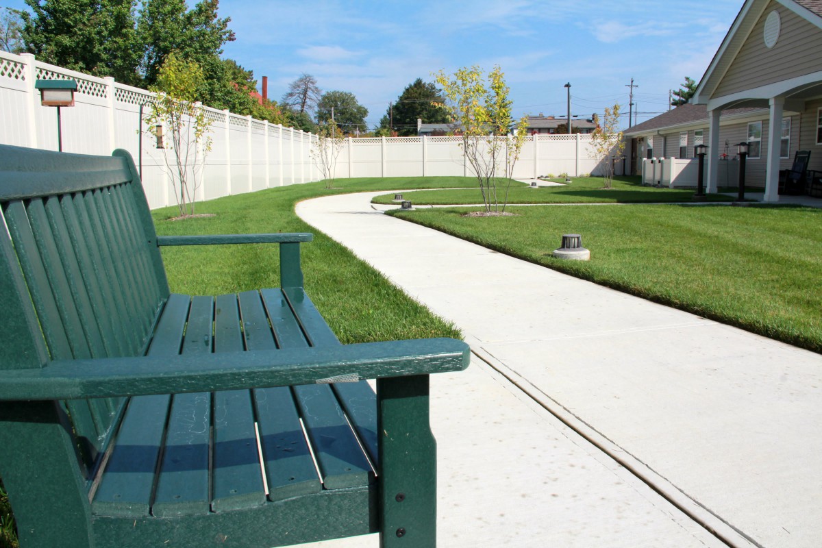 A peaceful park scene with a green wooden bench beside a paved pathway surrounded by grass.